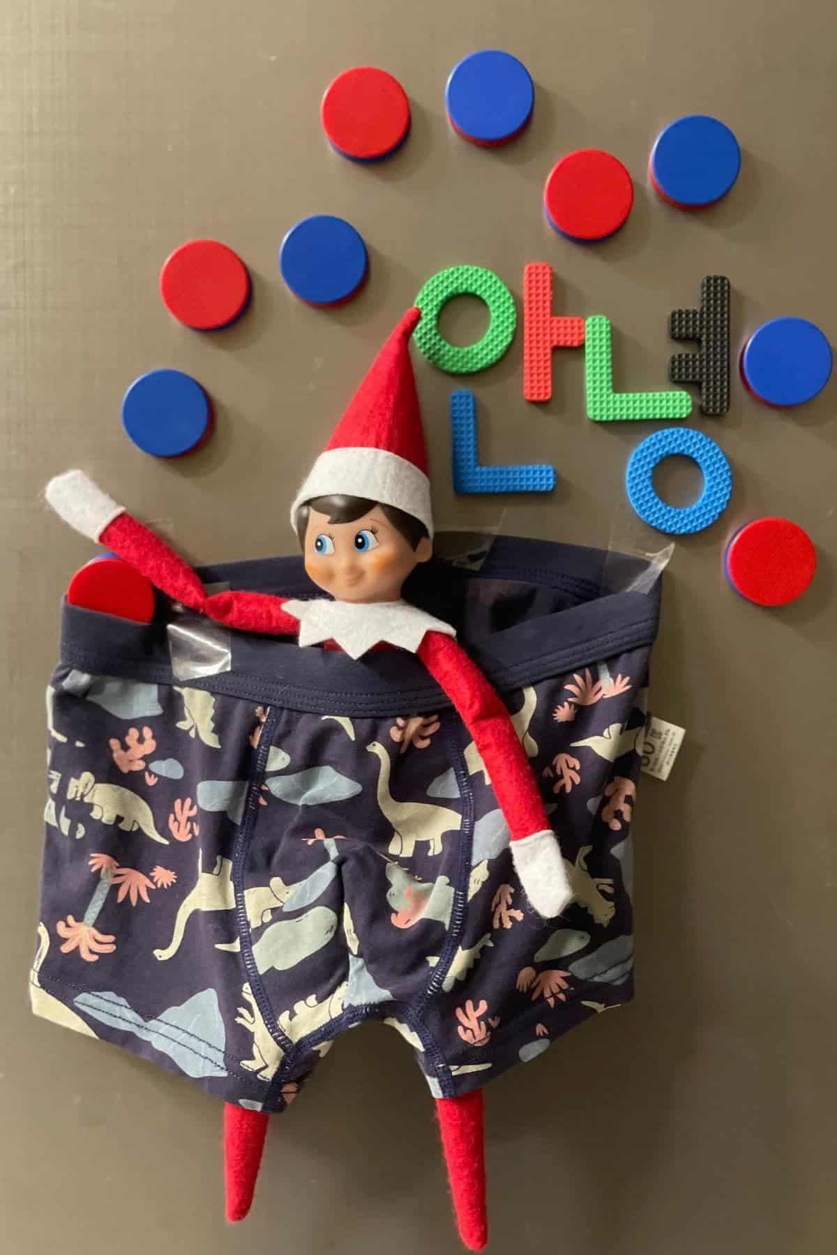 A toy elf is inside kids' dinosaur print underwear, surrounded by red and blue discs and foam letters spelling "Hello" on a gray surface.