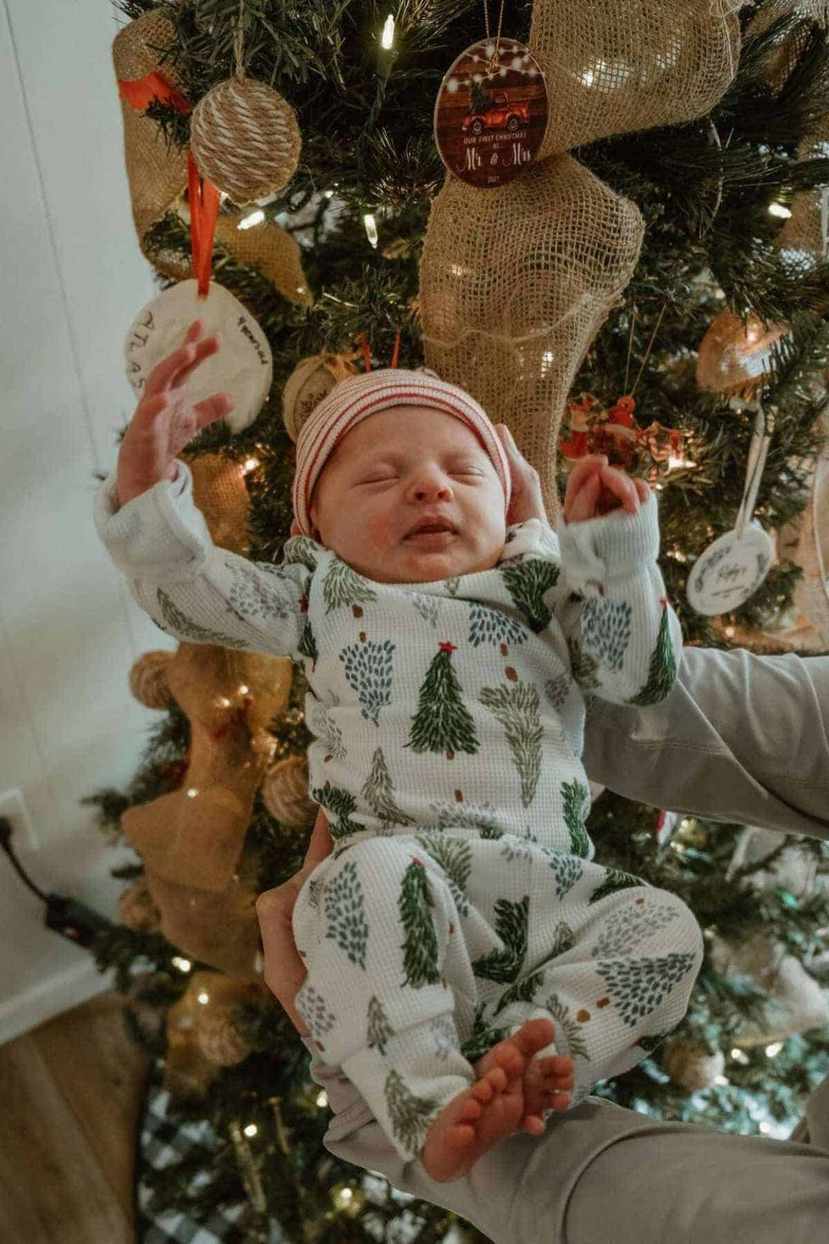 A baby in a holiday-themed onesie and hat is held up in front of a decorated Christmas tree with lights and ornaments.