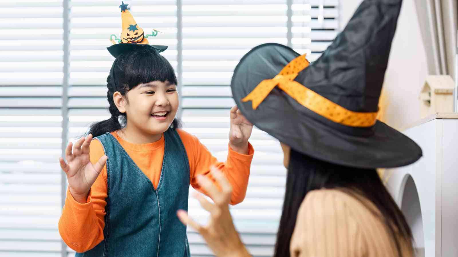 A child and an adult wearing Halloween hats and costumes face each other indoors, raising their hands as if pretending to be spooky.