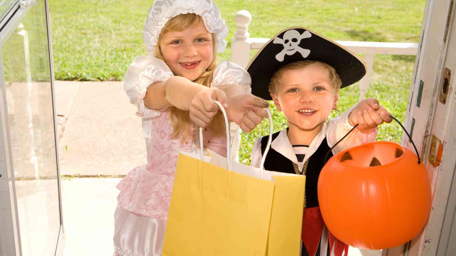 Two children in costume, one as a pirate and one as a princess, stand at a door holding out bags for trick-or-treating.