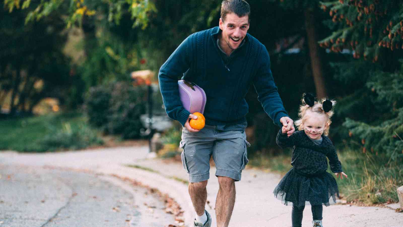 Adult holding a child’s hand while walking on a sidewalk; the child is dressed in a black costume, and the adult carries a pumpkin container and a helmet.