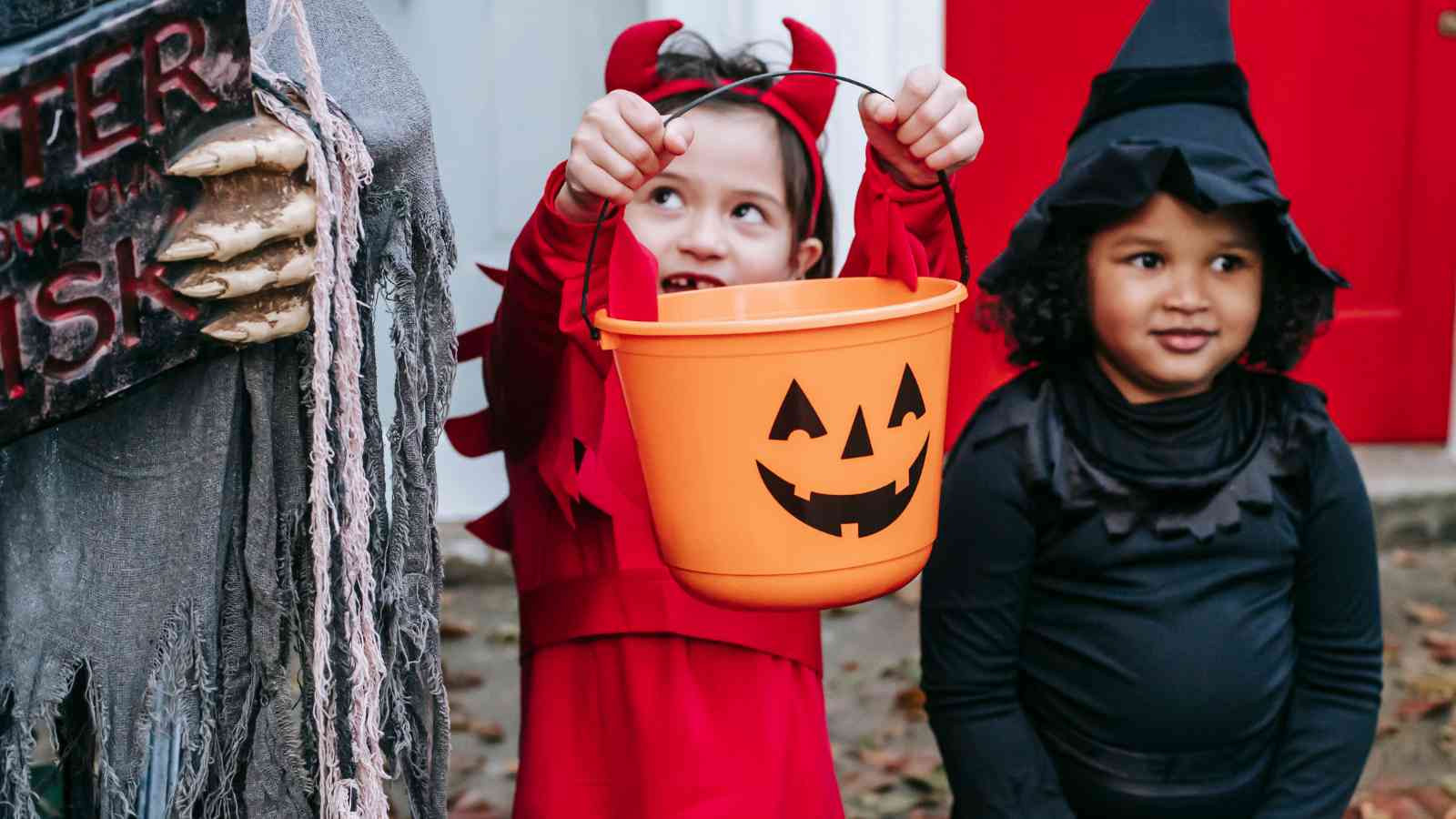 Two children in Halloween costumes, one dressed as a devil holding an orange pumpkin bucket, and the other dressed as a witch, standing outdoors.