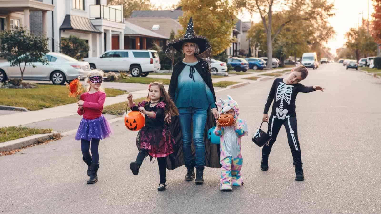 A group of children in Halloween costumes and an adult in a witch hat walk together on a suburban street during daylight.