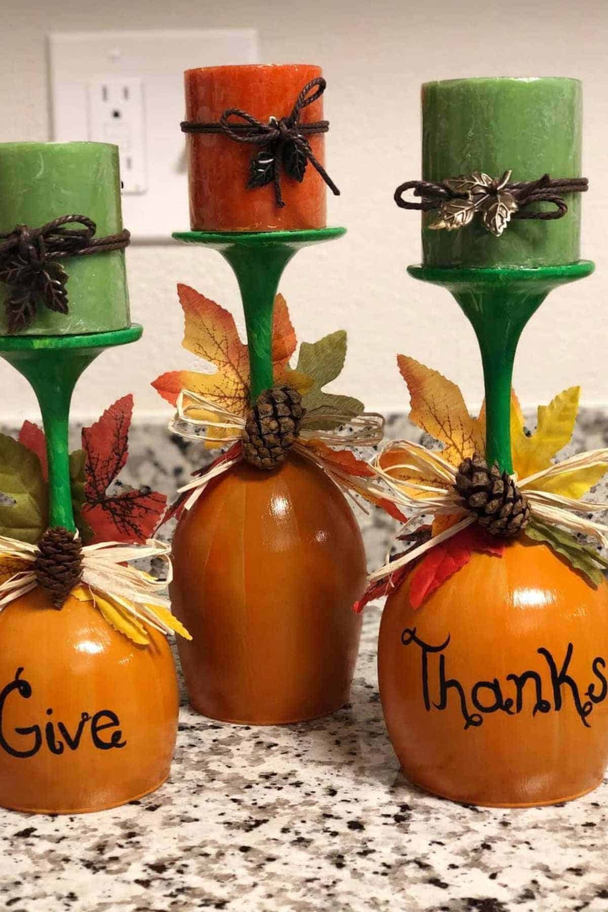 Three decorative candle holders made from upside-down wine glasses with fall leaves, pinecones, and the words "Give Thanks" written on the bases.