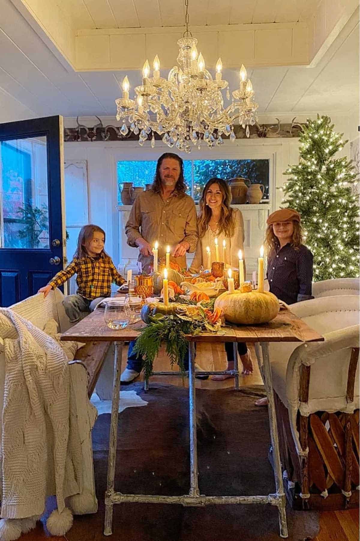 A family of four stands around a dining table set with candles and autumn decorations in a cozy, warmly lit room with a chandelier and a decorated Christmas tree.