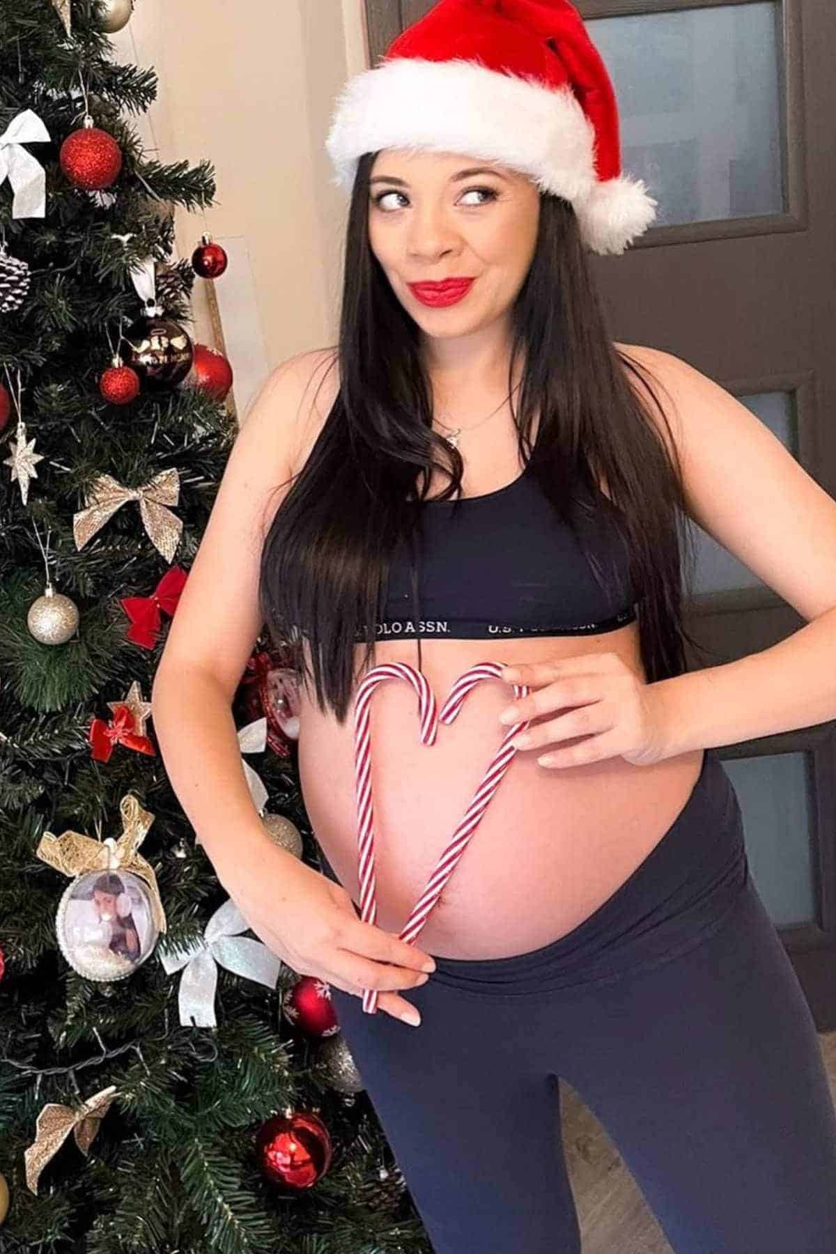 A pregnant woman in a Santa hat holds two candy canes shaped like a heart in front of her belly, standing next to a decorated Christmas tree.