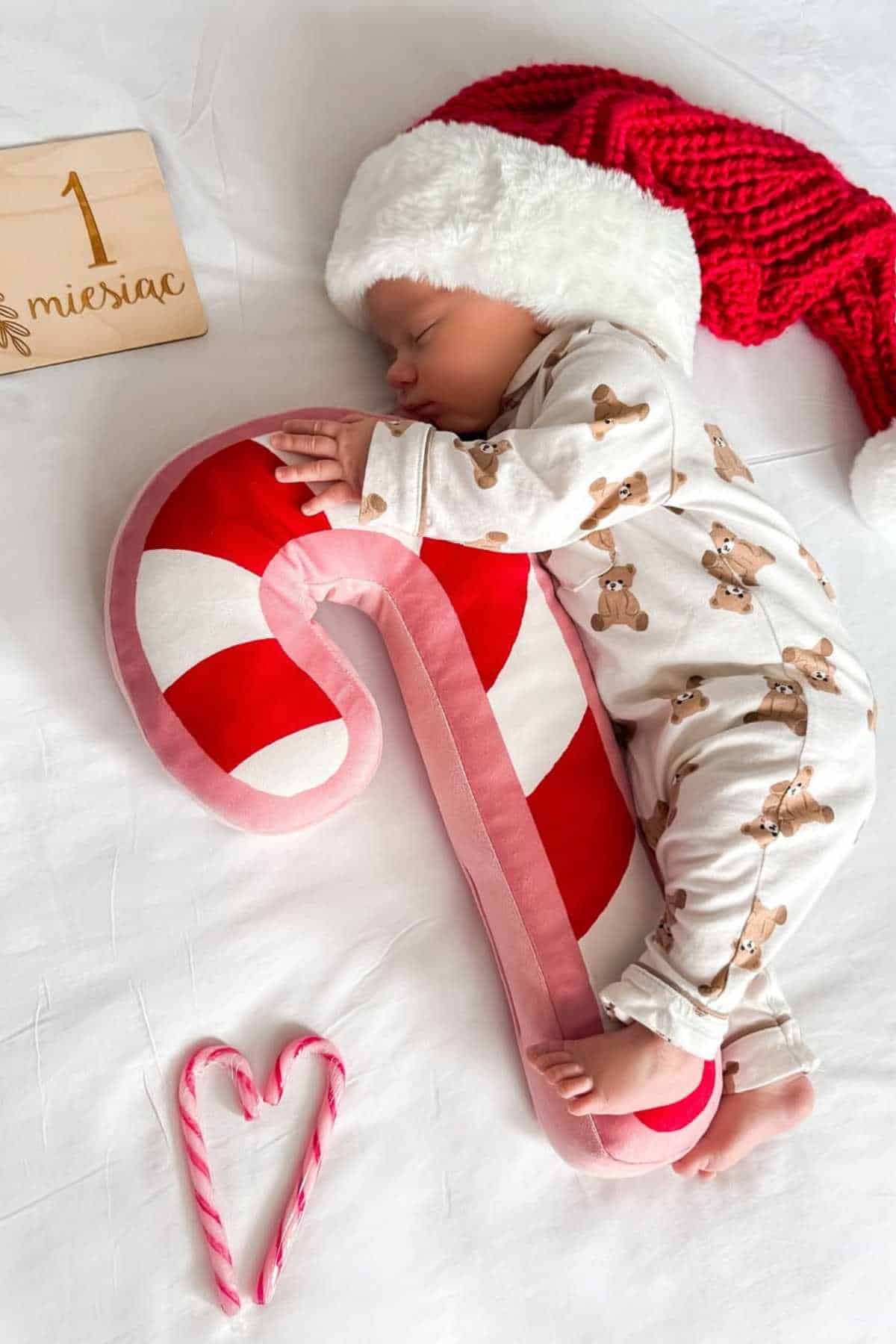 A baby in a Santa hat sleeps on a bed, hugging a large plush candy cane. The baby wears pajamas with gingerbread men and a "1 miesiąc" sign is nearby. Two candy canes form a heart shape.