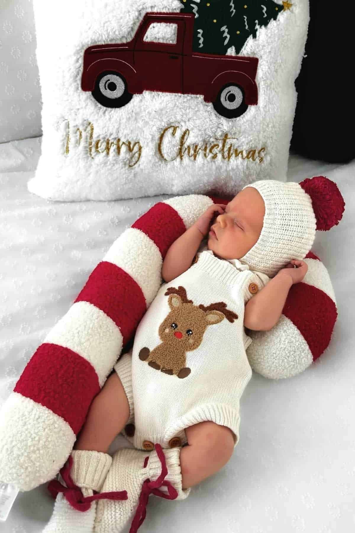 Newborn dressed in a reindeer romper and knit hat, lying on a red and white candy cane pillow, with a "Merry Christmas" cushion in the background.