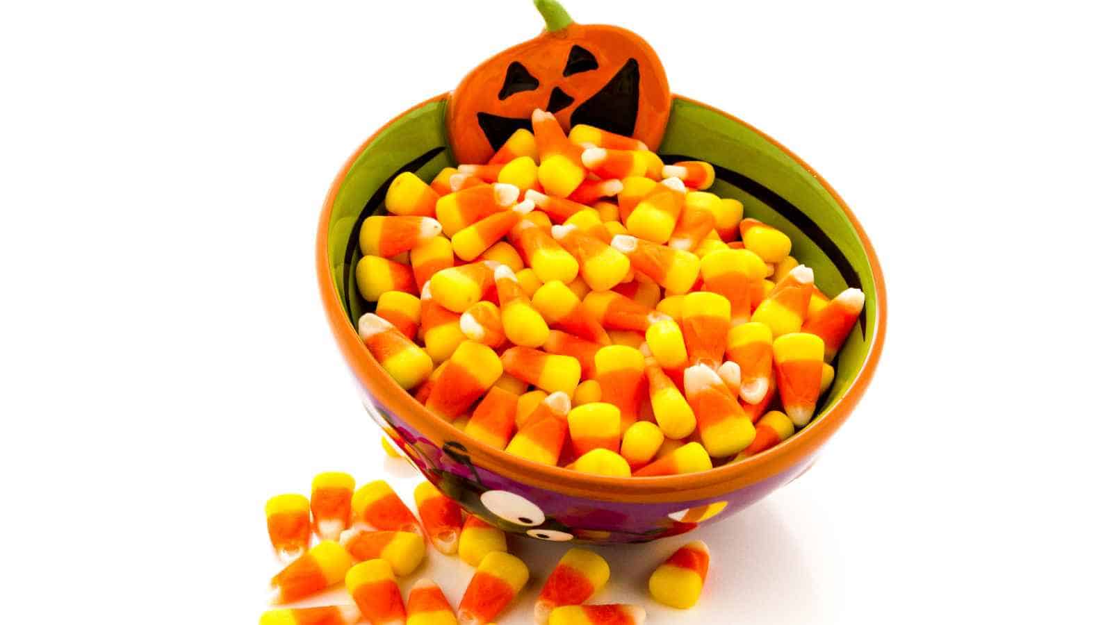 A bowl filled with candy corn sits on a white surface, with a small pumpkin decoration attached to the bowl and some candy corn pieces scattered outside.