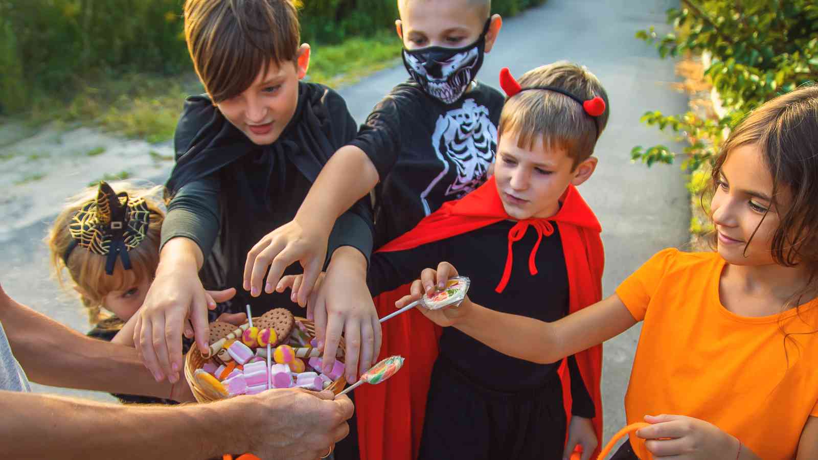 Five children in Halloween costumes reach for candy from a basket held by an adult outdoors.