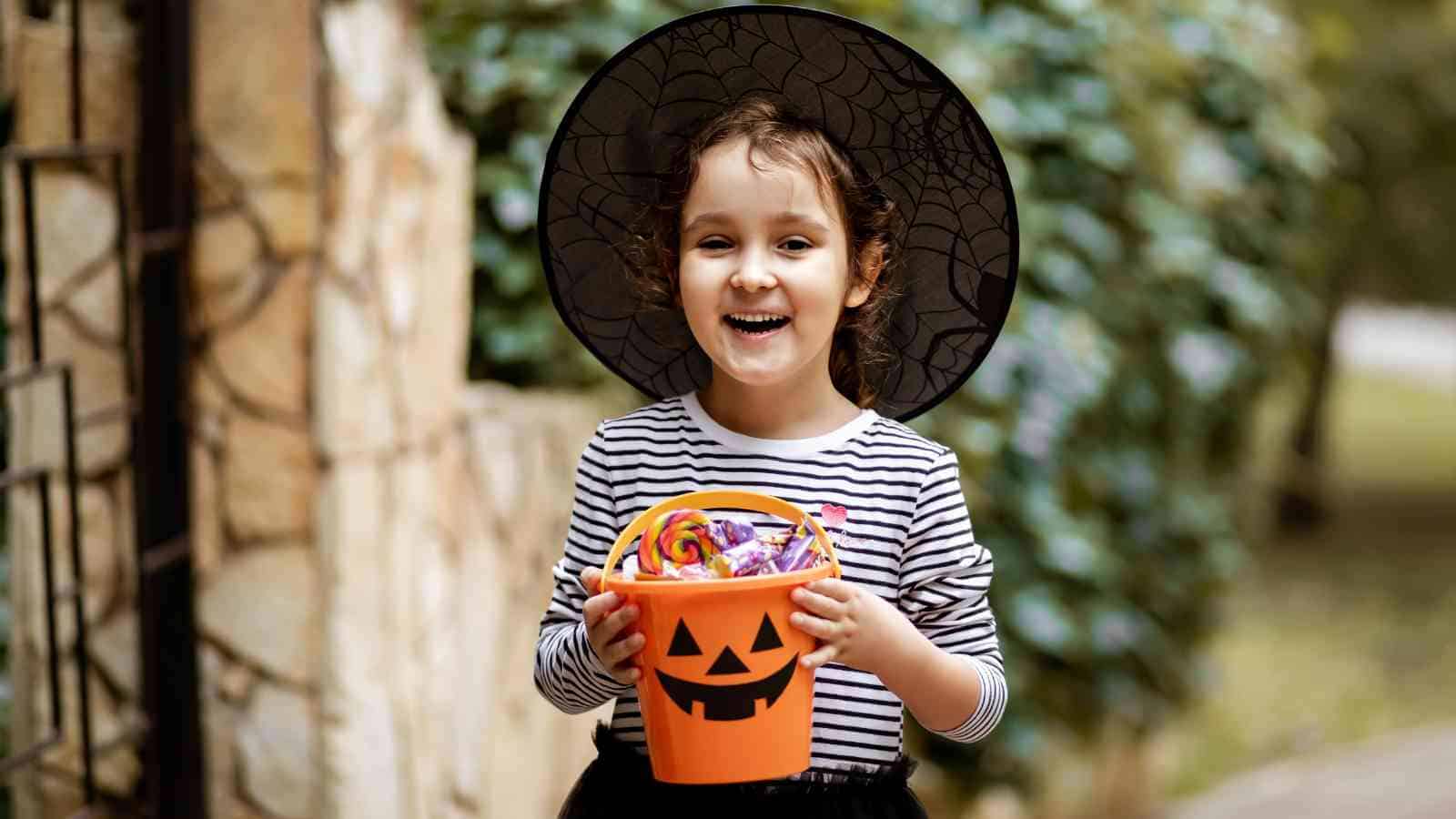 Child wearing a witch hat and striped shirt smiles while holding an orange pumpkin bucket filled with Halloween candy outdoors.