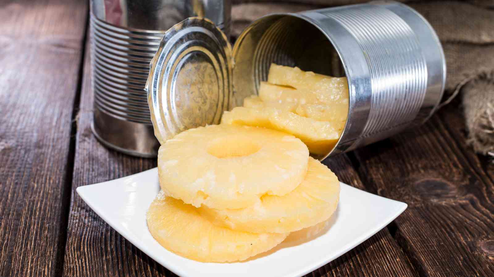 Slices of canned pineapple are arranged on a white plate, with an open can and lid in the background on a wooden surface.