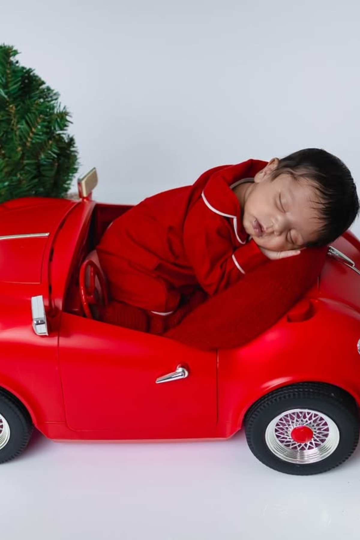 A baby dressed in red pajamas sleeps inside a small red toy car, with a green plant partially visible in the background.