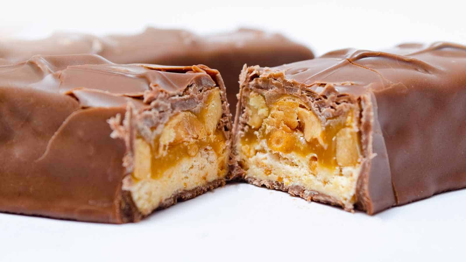 A chocolate bar cut in half reveals layers of caramel, nougat, and peanuts inside.