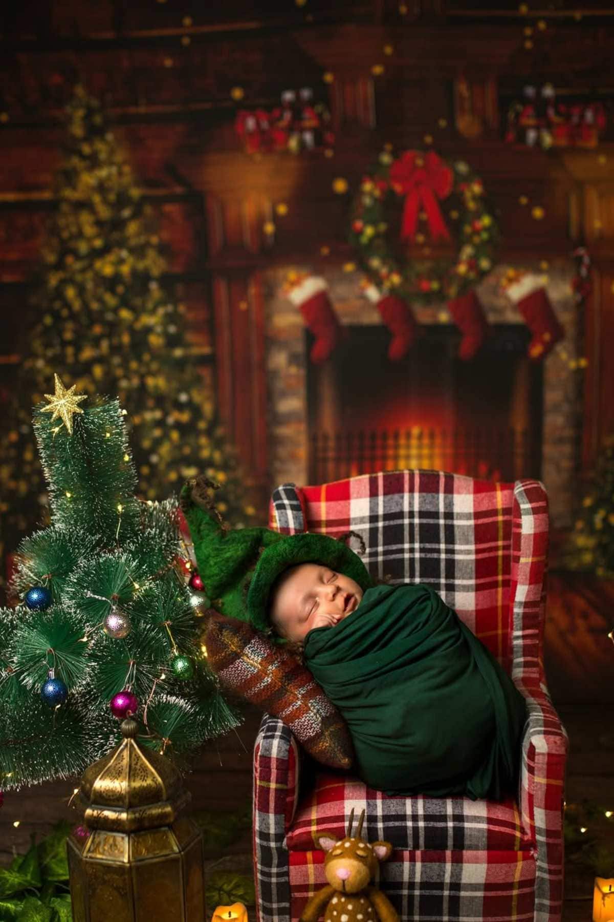 A newborn baby wrapped in a green blanket sleeps on a plaid chair beside a small decorated Christmas tree, with a festive fireplace and wreath in the background.