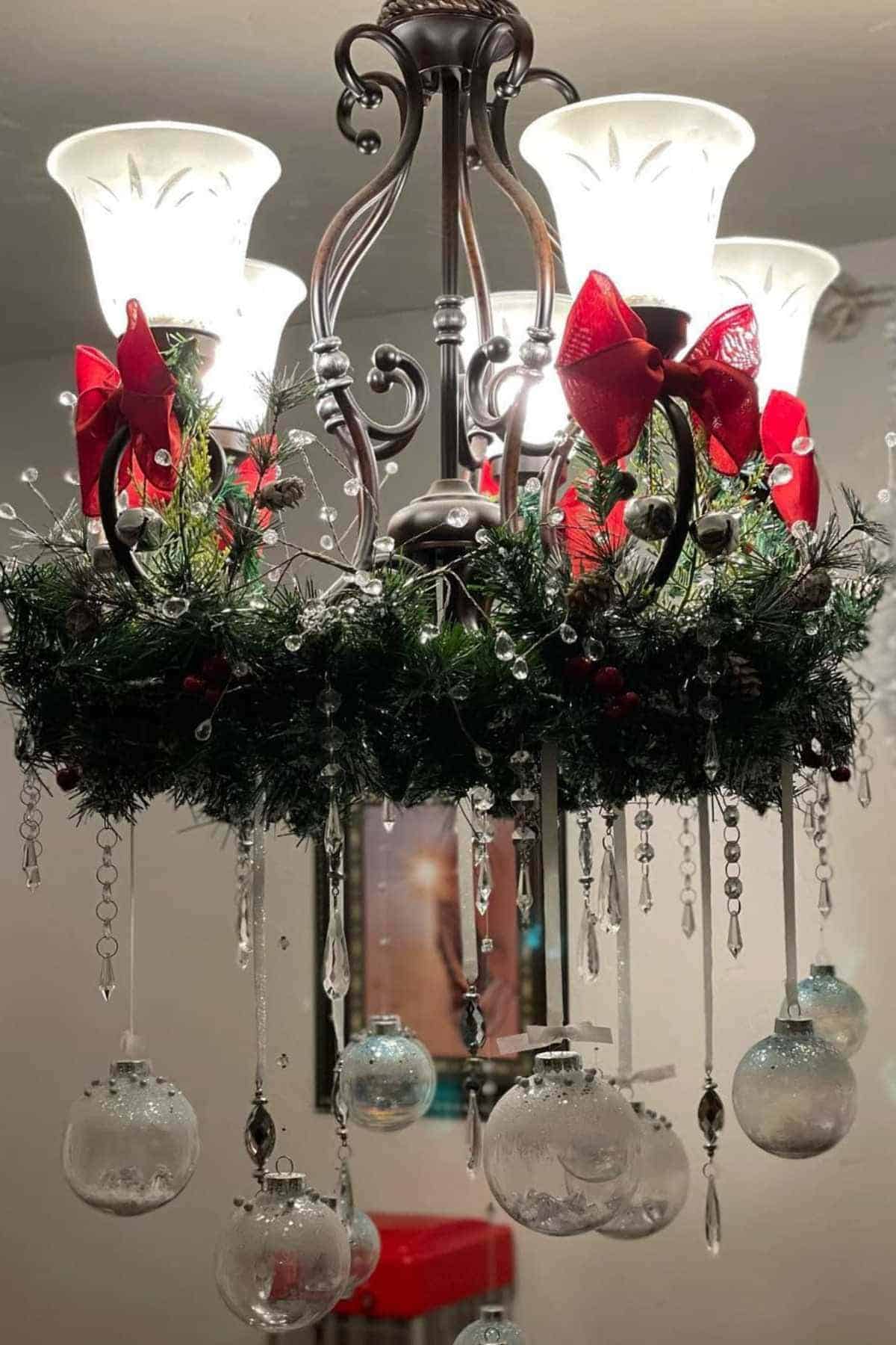 A chandelier decorated with greenery, red bows, hanging crystal ornaments, and clear glass baubles, illuminated by several lights.