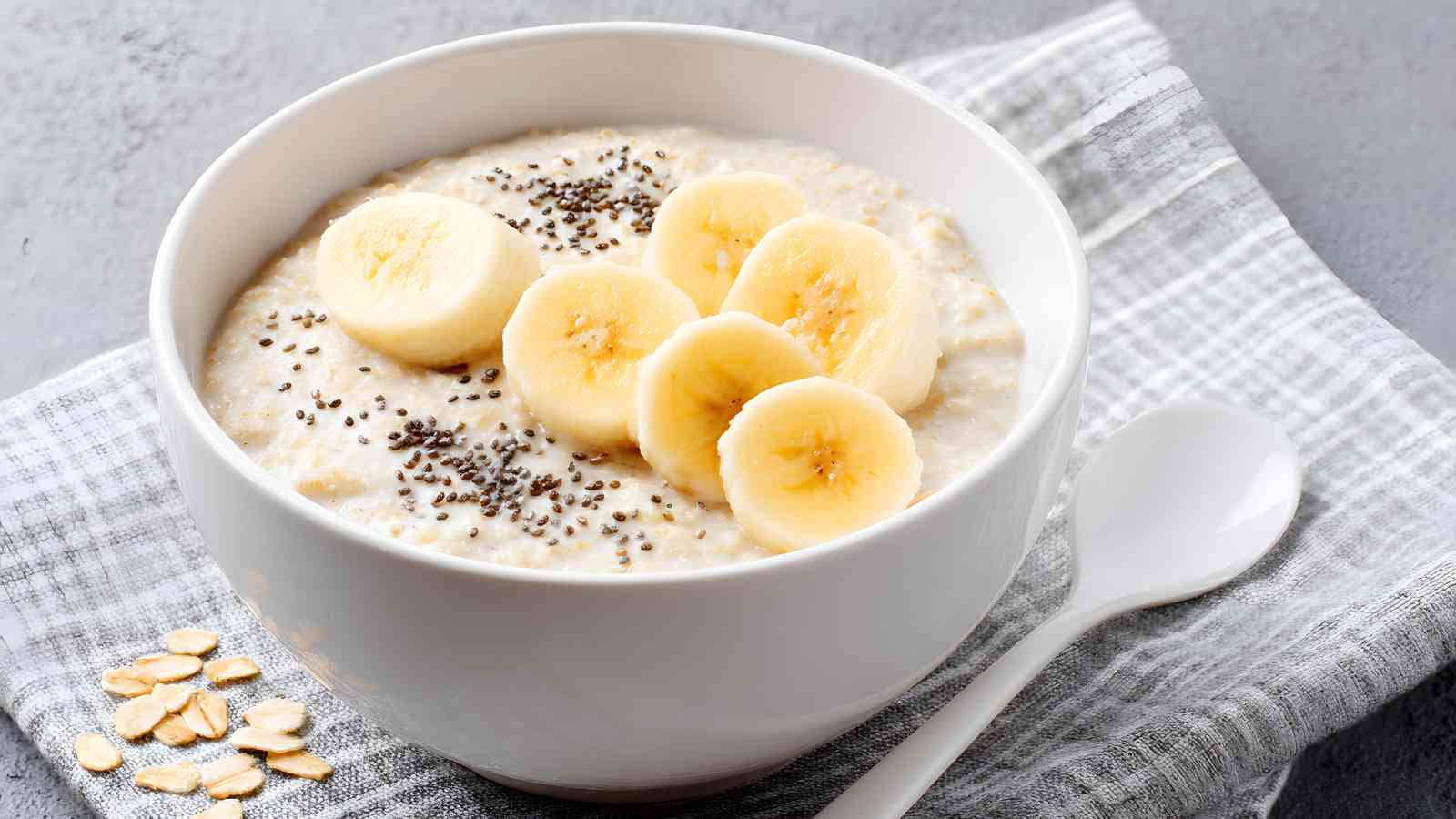 A white bowl of oatmeal topped with banana slices and chia seeds sits on a gray plaid cloth beside a white spoon.