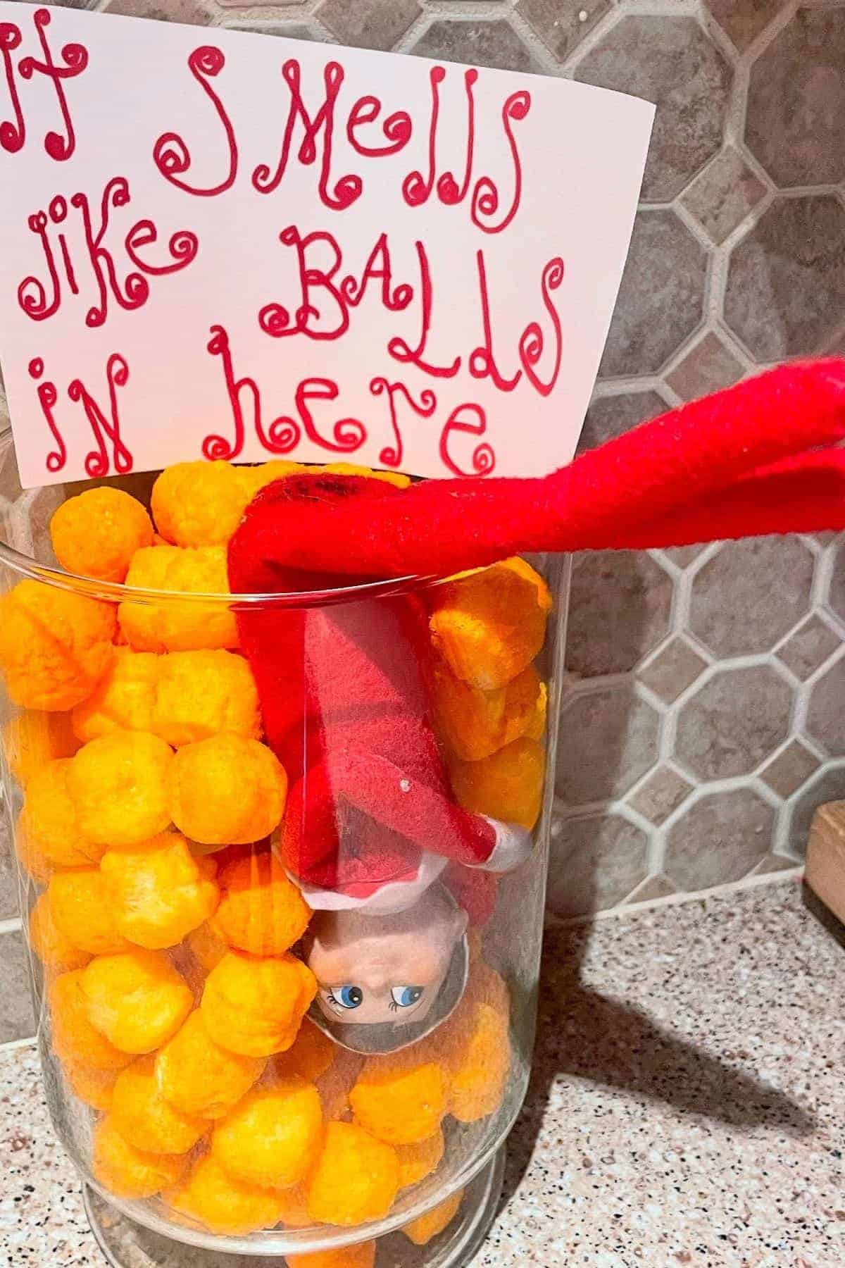 A toy elf is upside down in a glass jar filled with cheese balls. A handwritten sign behind it says, "It smells like balls in here.