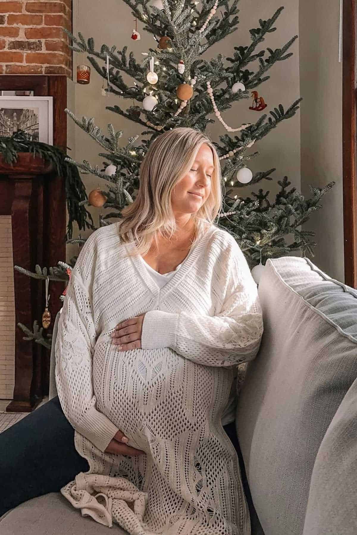 A pregnant woman in a white sweater sits on a couch by a decorated Christmas tree, resting her hands on her belly and looking peacefully out the window.