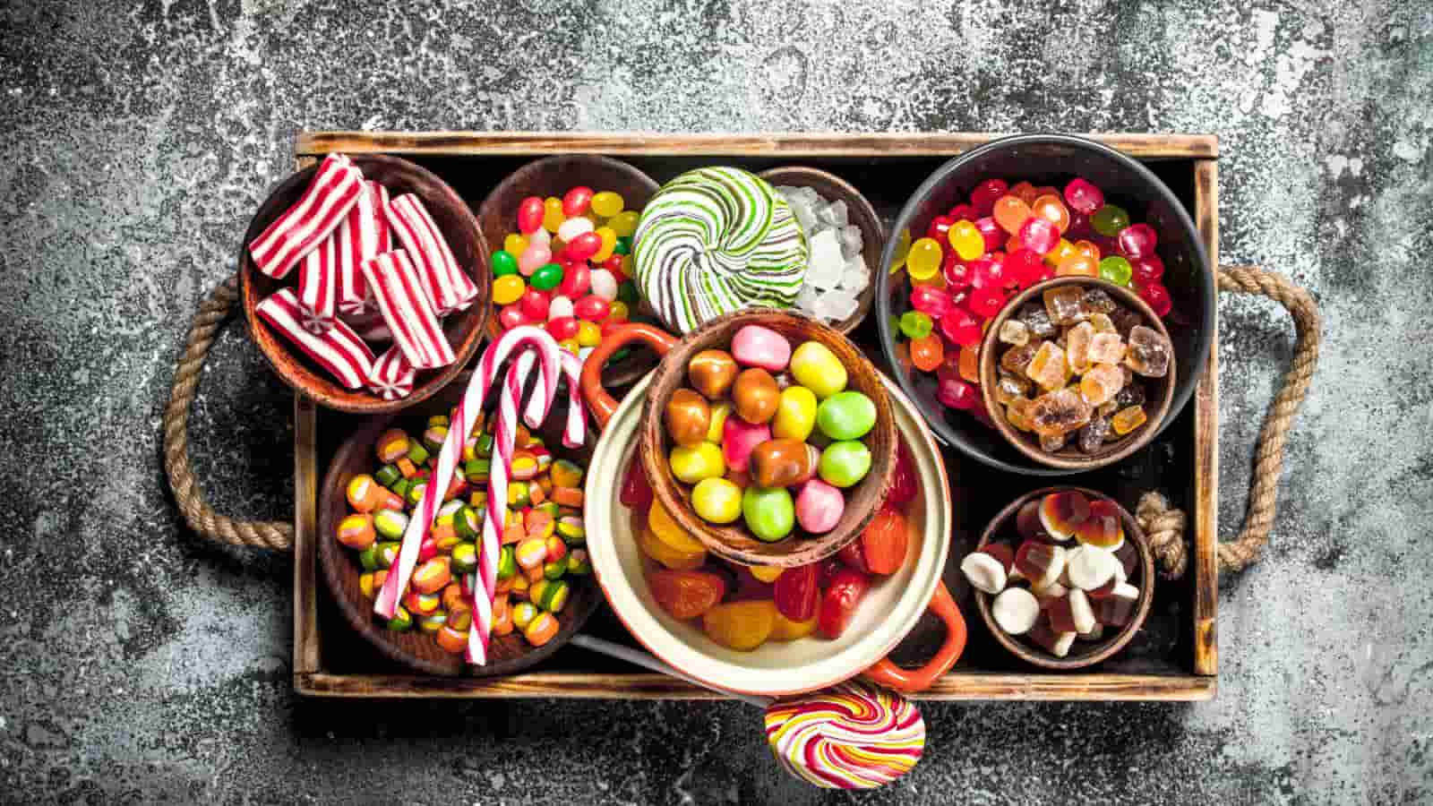 A wooden tray holding various bowls filled with assorted colorful candies, including jelly beans, candy canes, and hard candies, on a textured surface.