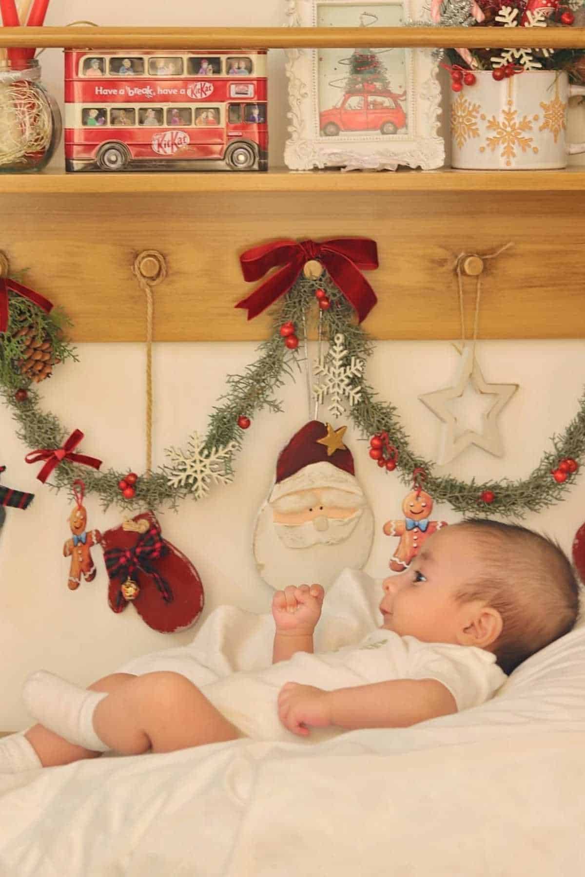 A baby lies on a bed under holiday decorations, including garlands, red bows, stockings, and a Santa ornament, with a shelf displaying a red bus and framed pictures above.