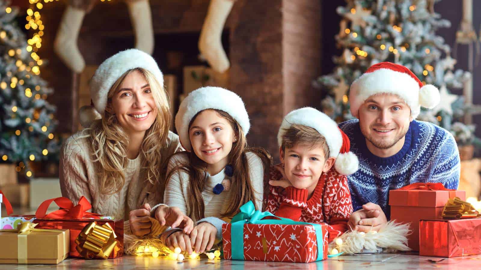 A family of four in Santa hats smiles together in front of a Christmas tree, surrounded by wrapped gifts and festive decorations.