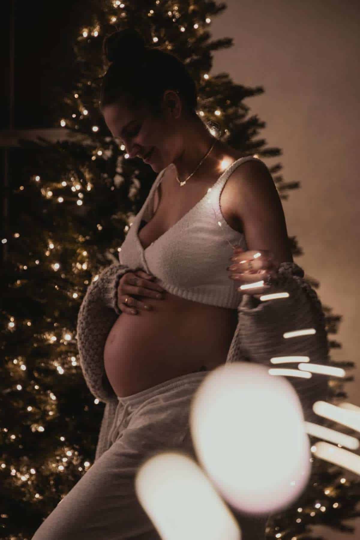 Pregnant woman in cozy clothes stands in front of a lit Christmas tree, holding her belly and surrounded by string lights.