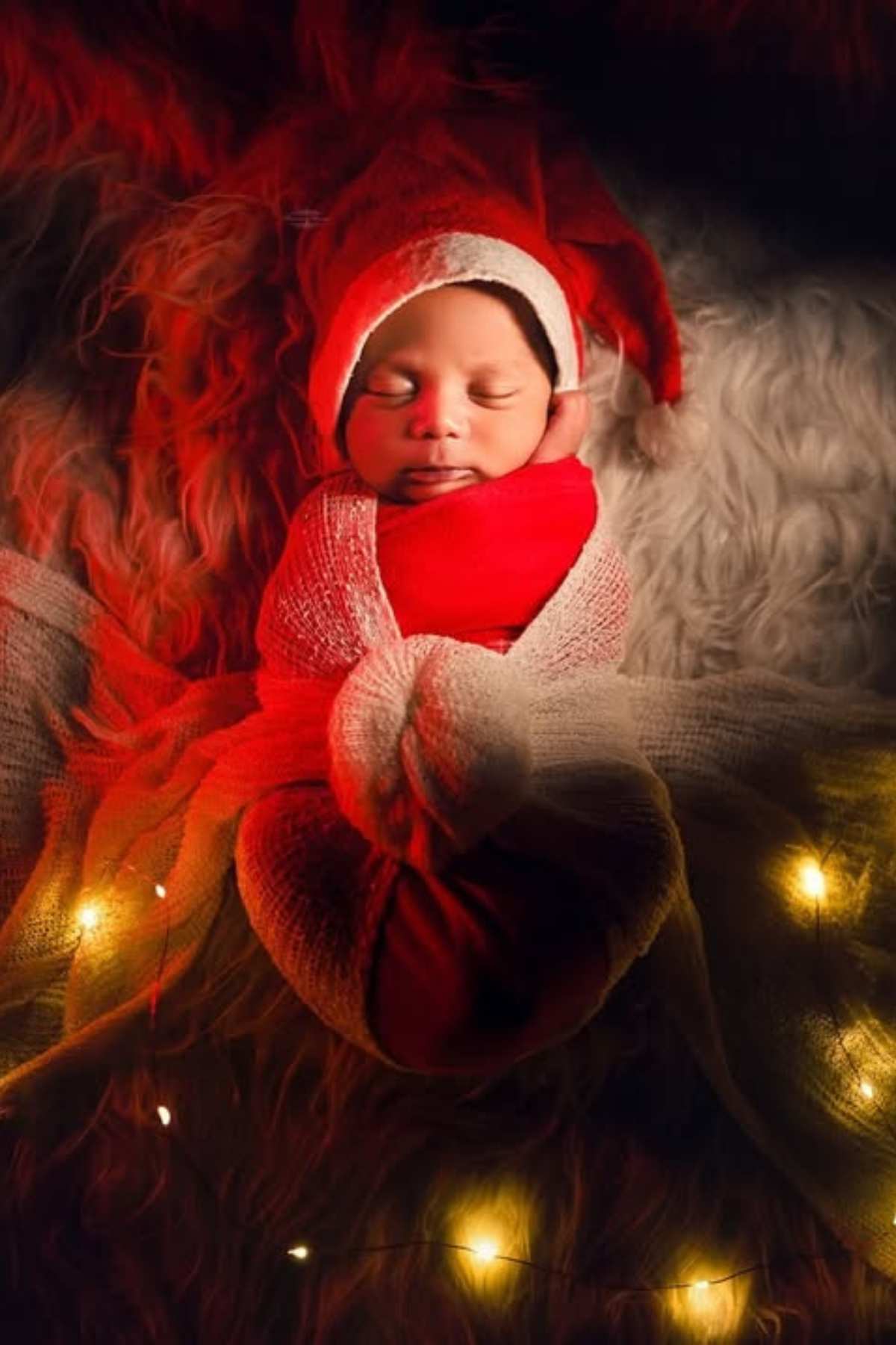 A newborn baby wrapped in red and white cloth with a Santa hat, lying on a furry surface, surrounded by warm fairy lights.