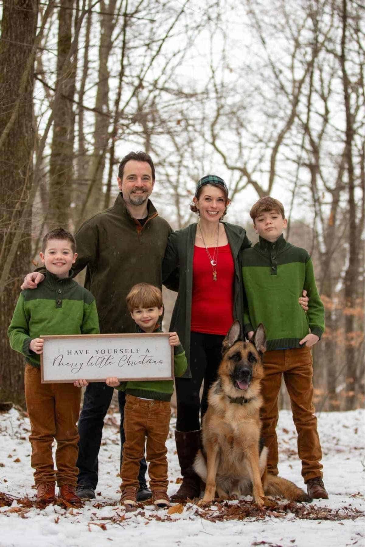 A family of five and a German Shepherd dog pose outdoors in a snowy forest. The children hold a sign that reads "Have Yourself a Merry Little Christmas.