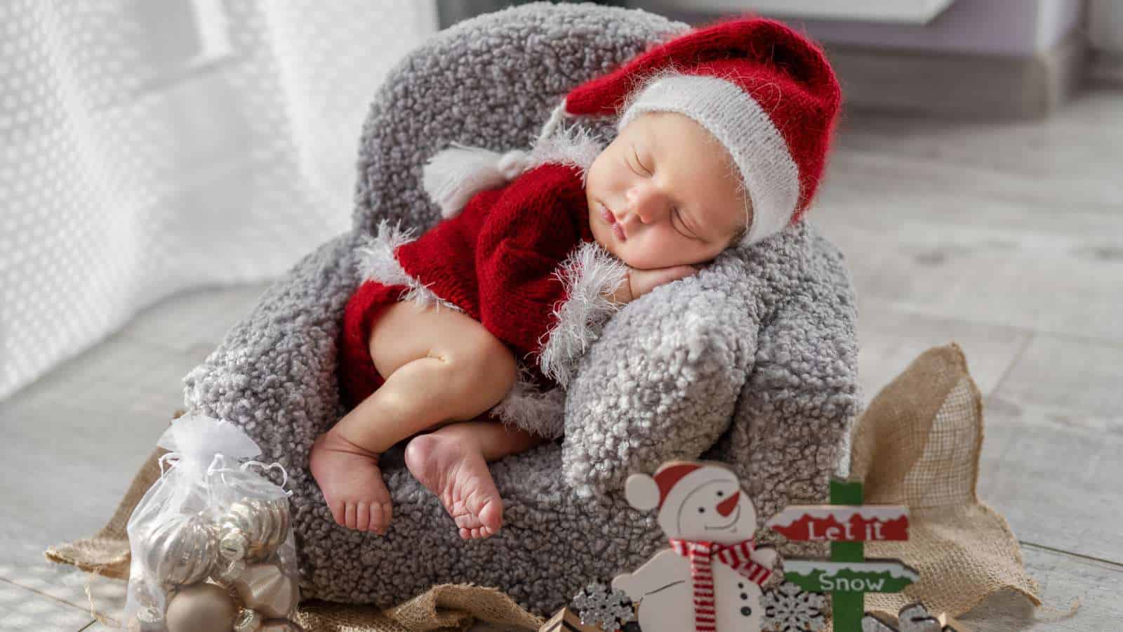 A newborn baby dressed in a red and white Santa outfit sleeps on a gray plush chair next to Christmas decorations and a snowman sign.