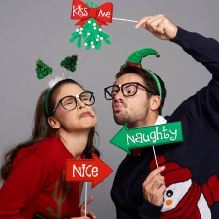 Two people wearing Christmas sweaters and headbands pose playfully; one holds a "Nice" sign and the other a "Naughty" sign with a "Kiss Me" mistletoe prop over their heads.