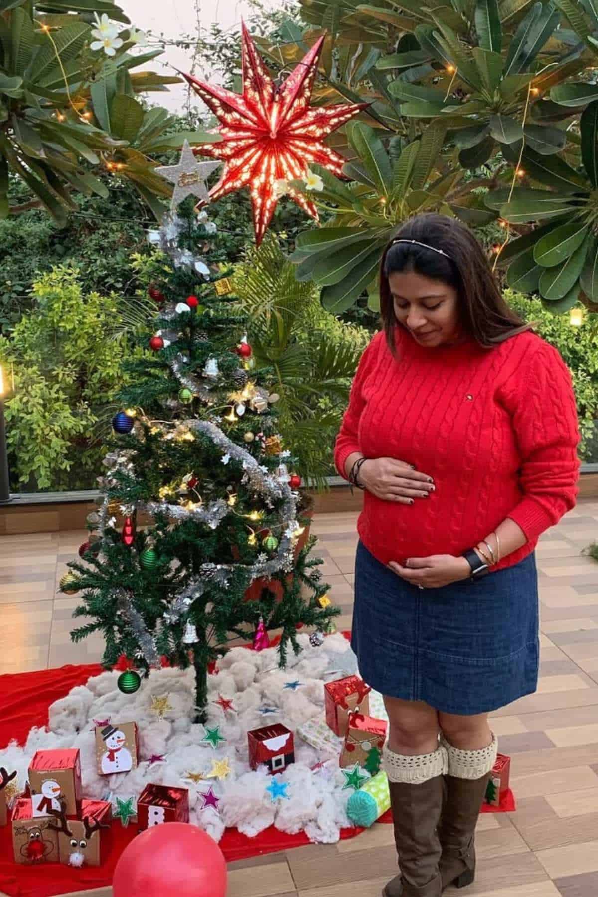 A woman in a red sweater stands by a decorated Christmas tree, holding her belly and looking down, with wrapped gifts and festive decorations around her.