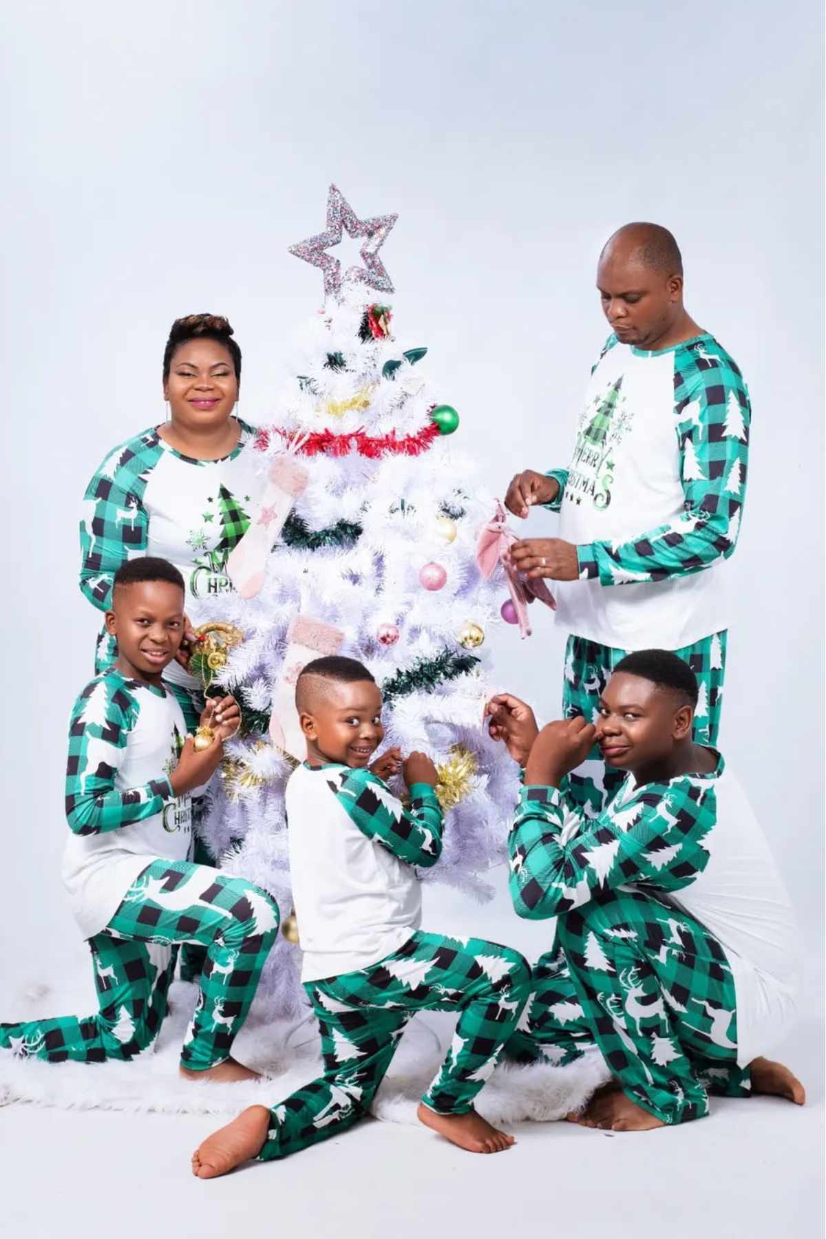A family in matching green and white pajamas decorates a white Christmas tree together against a plain background.