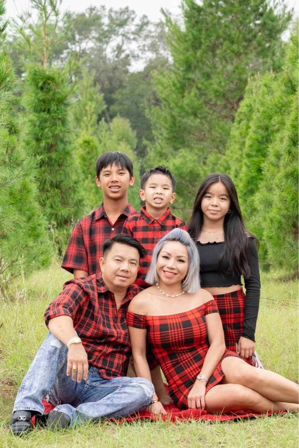 A family of five poses outdoors among green trees, all wearing coordinated red and black plaid outfits and smiling at the camera.