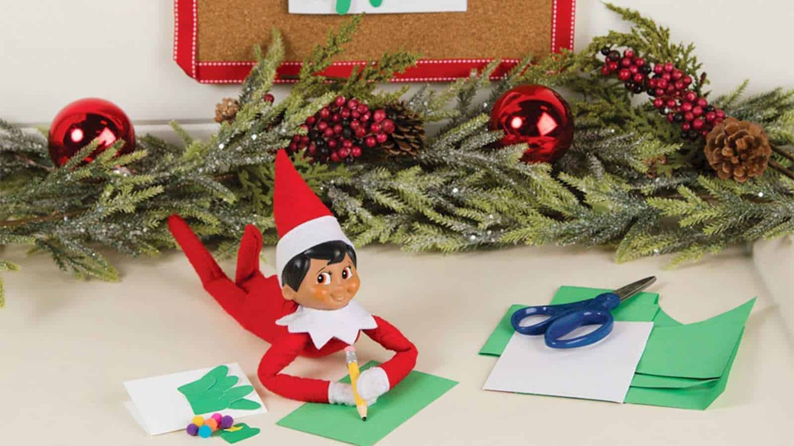 A toy elf lies on a table, drawing on green paper with a pencil. Scissors, paper, and decorative greenery with red ornaments are nearby.