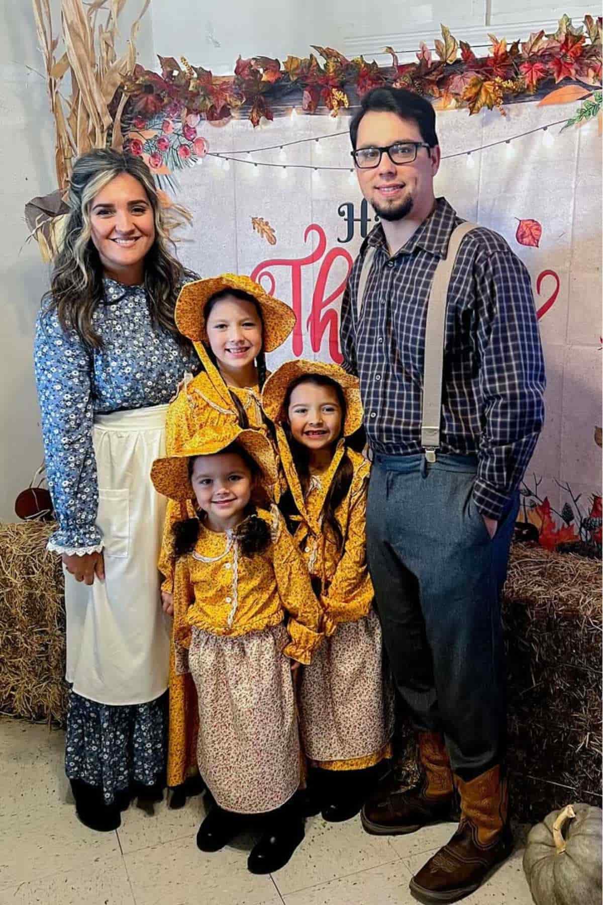 A family poses together in pioneer-style clothing, with three young girls in matching bonnets and dresses, in front of a fall-themed backdrop.