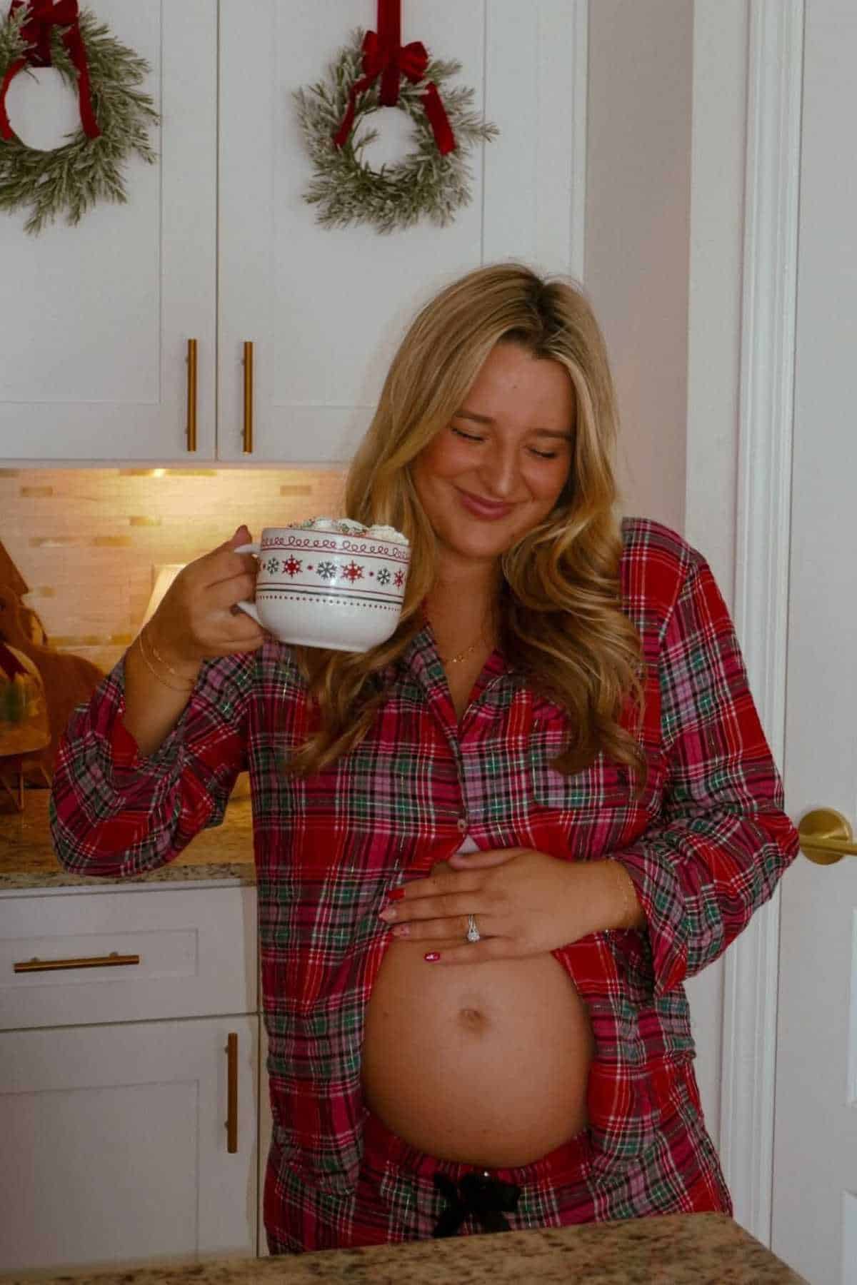 A woman in red plaid pajamas holds a mug and cradles her pregnant belly, standing in a kitchen with Christmas wreaths on white cabinets.