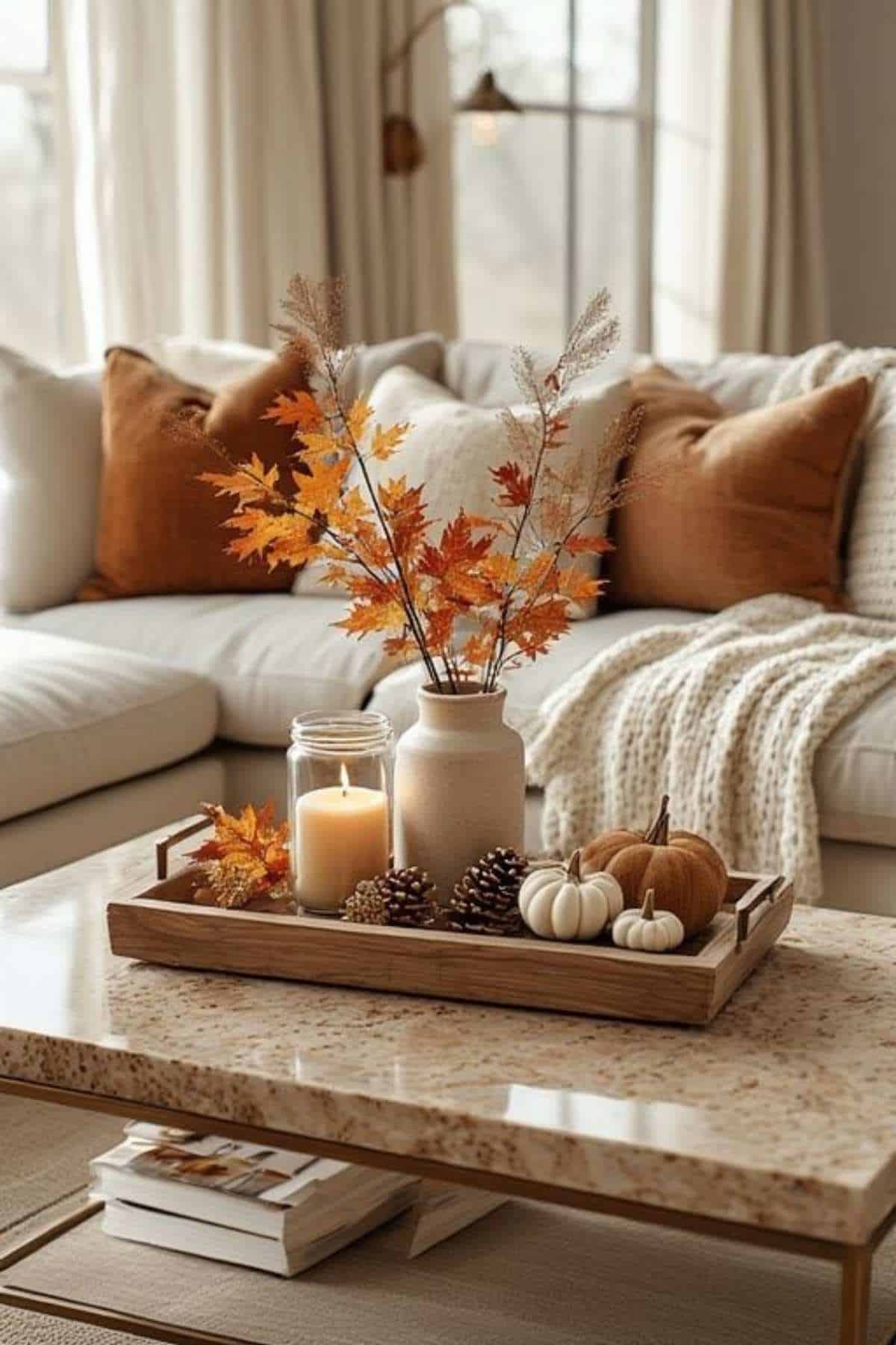 A coffee table displays autumn decor with a vase of orange leaves, a lit candle, pinecones, and small pumpkins; a beige sofa with brown pillows is in the background.