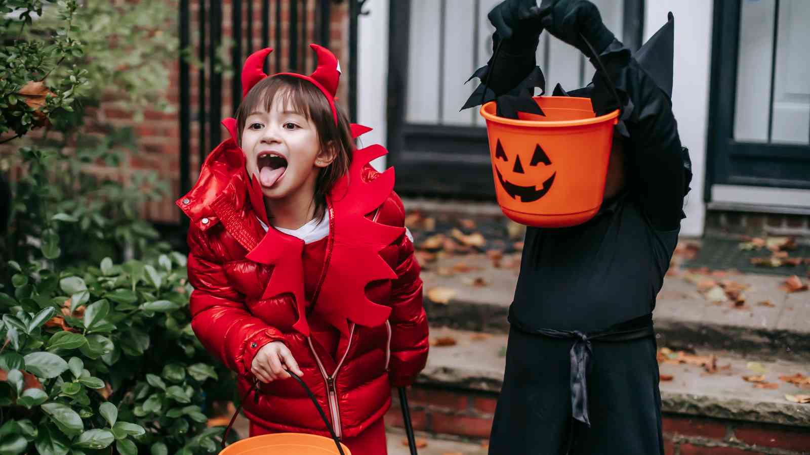 Two children in Halloween costumes stand outside; one dressed as a devil sticking out her tongue, and the other in black holding a pumpkin bucket over their face.