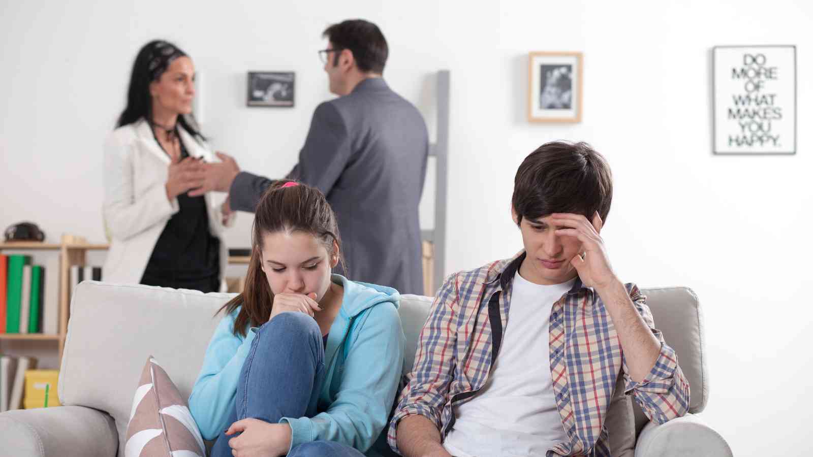 Two teenagers sit on a couch looking upset while two adults talk in the background of a living room.