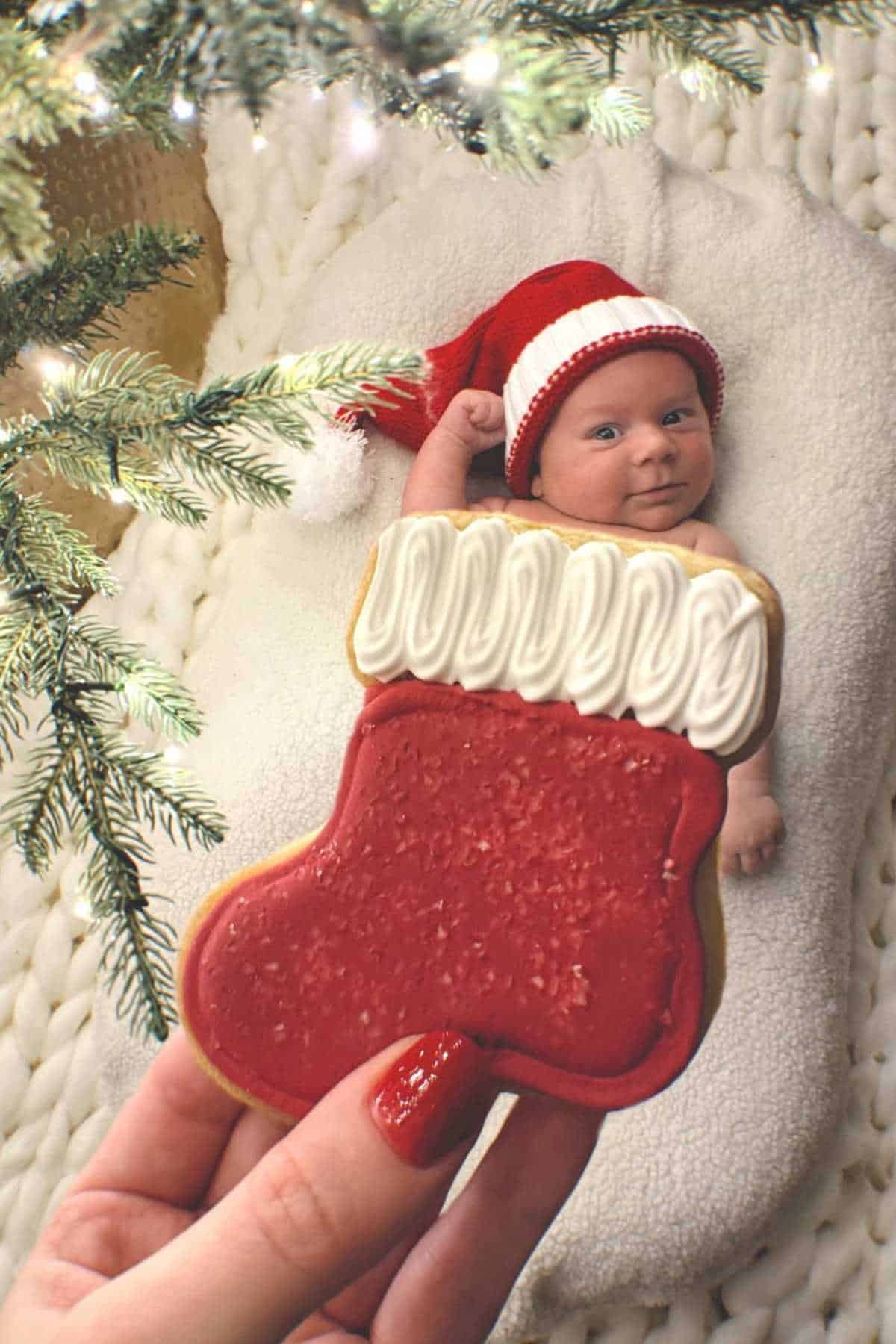 A baby in a red Santa hat lies on a blanket under a Christmas tree, with a hand holding a decorated stocking-shaped cookie in the foreground.