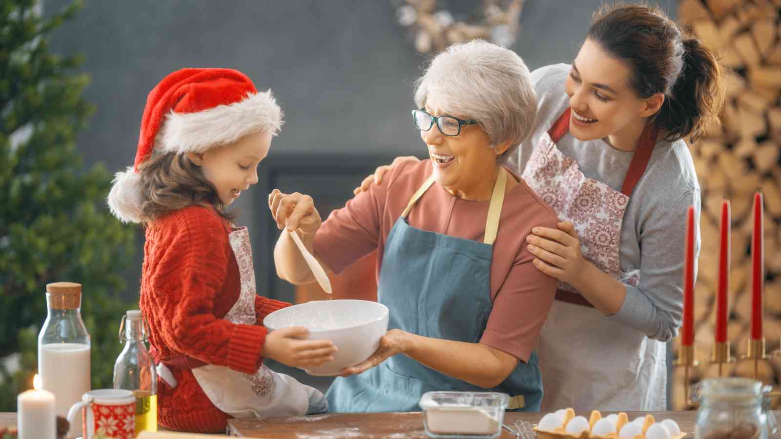 A child in a Santa hat, an older woman, and a younger woman mix ingredients in a bowl together at a kitchen table decorated for the holidays.