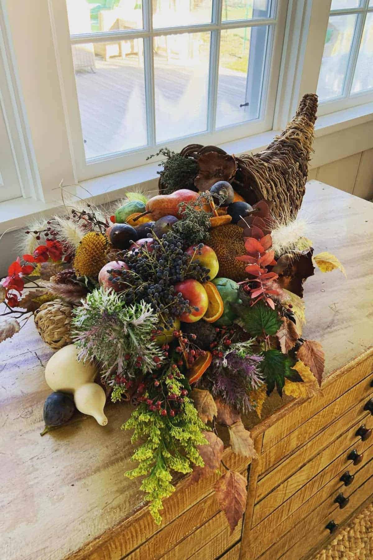 A woven cornucopia filled with assorted artificial fruits, vegetables, and autumn foliage sits on a wooden table in front of a window.