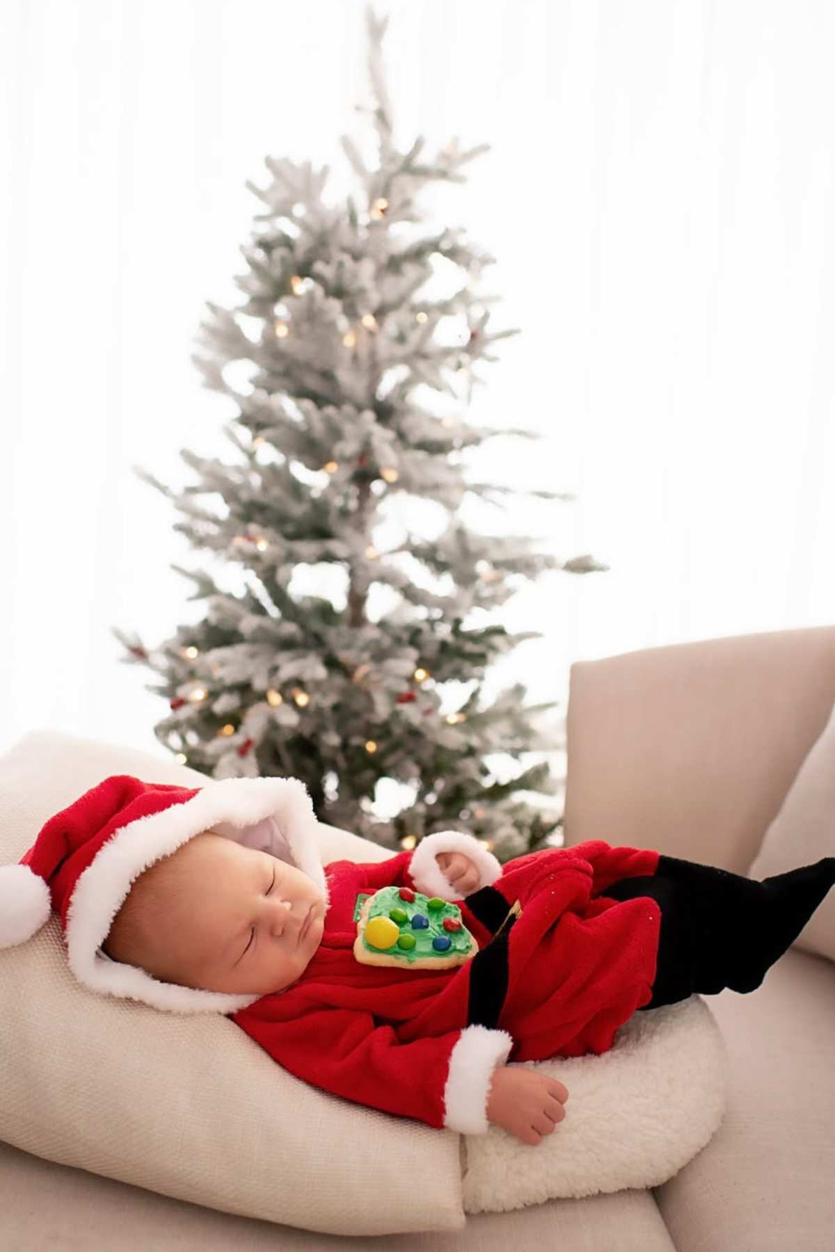 A baby dressed in a Santa outfit sleeps on a cushion, holding a toy, with a decorated Christmas tree in the background.