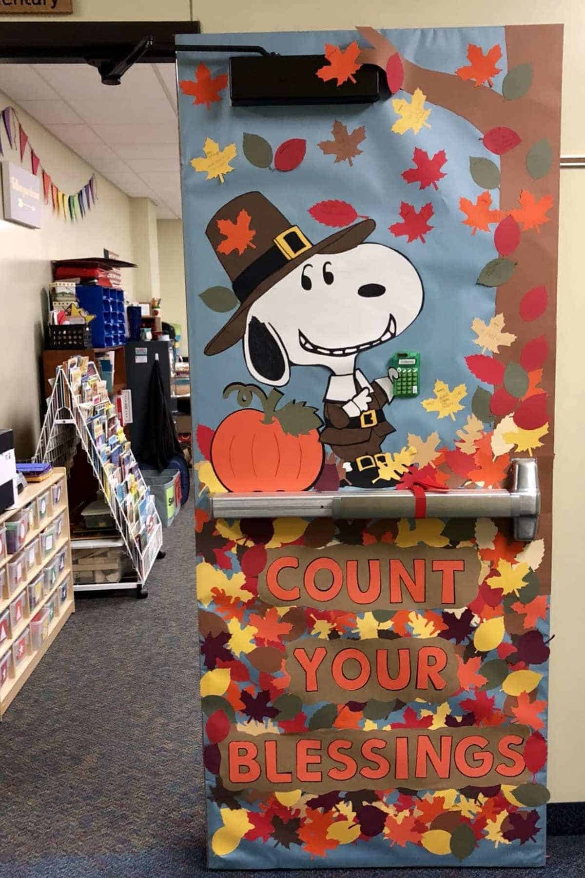 A classroom door decorated with a Snoopy character in a pilgrim hat, autumn leaves, a pumpkin, and the words "COUNT YOUR BLESSINGS" in large letters.