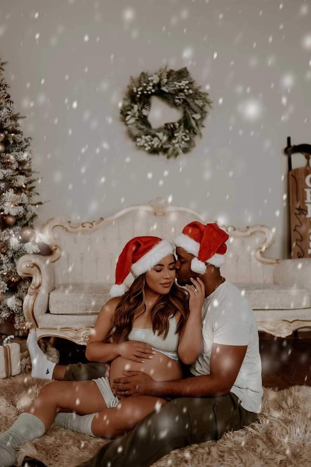 A couple wearing Santa hats sits on the floor by a Christmas tree and wreath; the woman is pregnant, and the man kisses her forehead while snow appears to fall indoors.