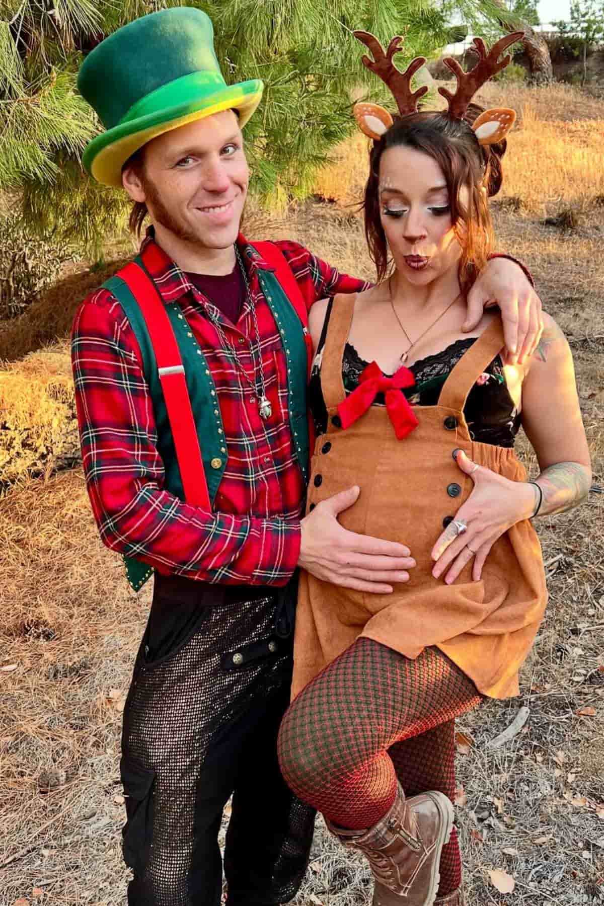 Two people in festive costumes pose outdoors; one is dressed as an elf and the other as a pregnant reindeer, with antlers and a red bow on the dress.
