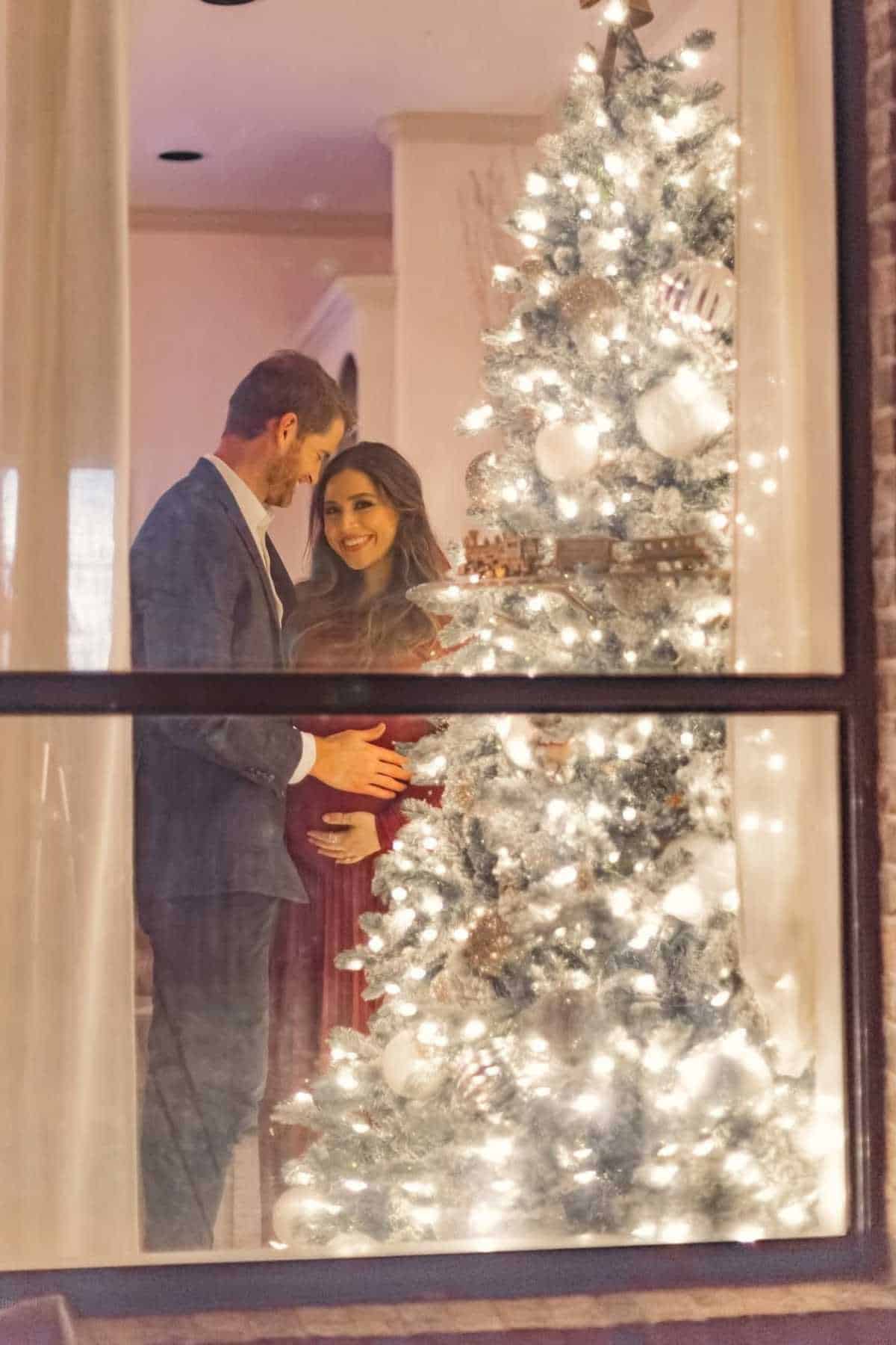 A man and a woman stand beside a decorated, lit Christmas tree indoors. The man touches the woman's pregnant belly as they both smile, seen through a window.