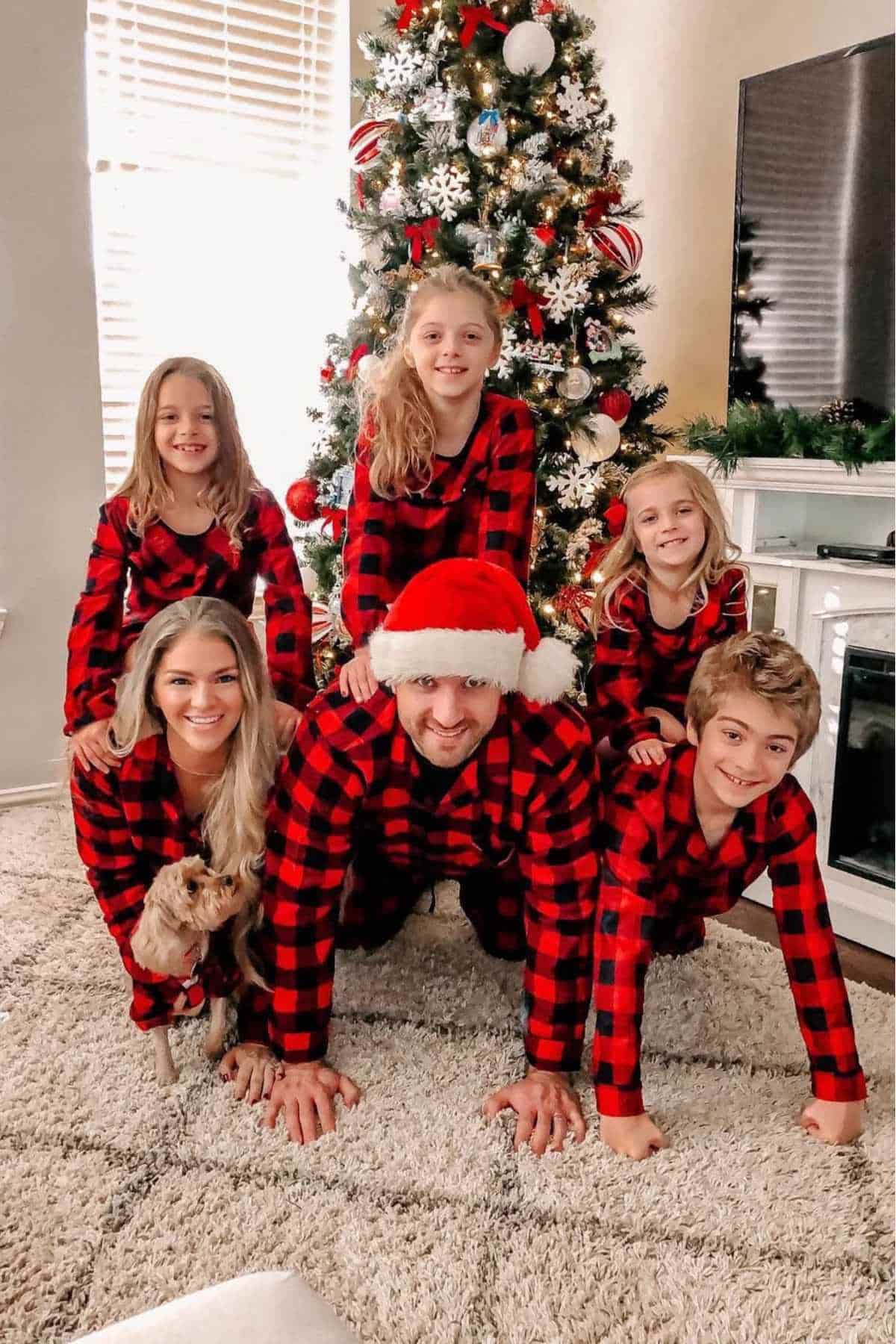 A family in matching red and black plaid pajamas poses in front of a decorated Christmas tree, with a small dog in the foreground.