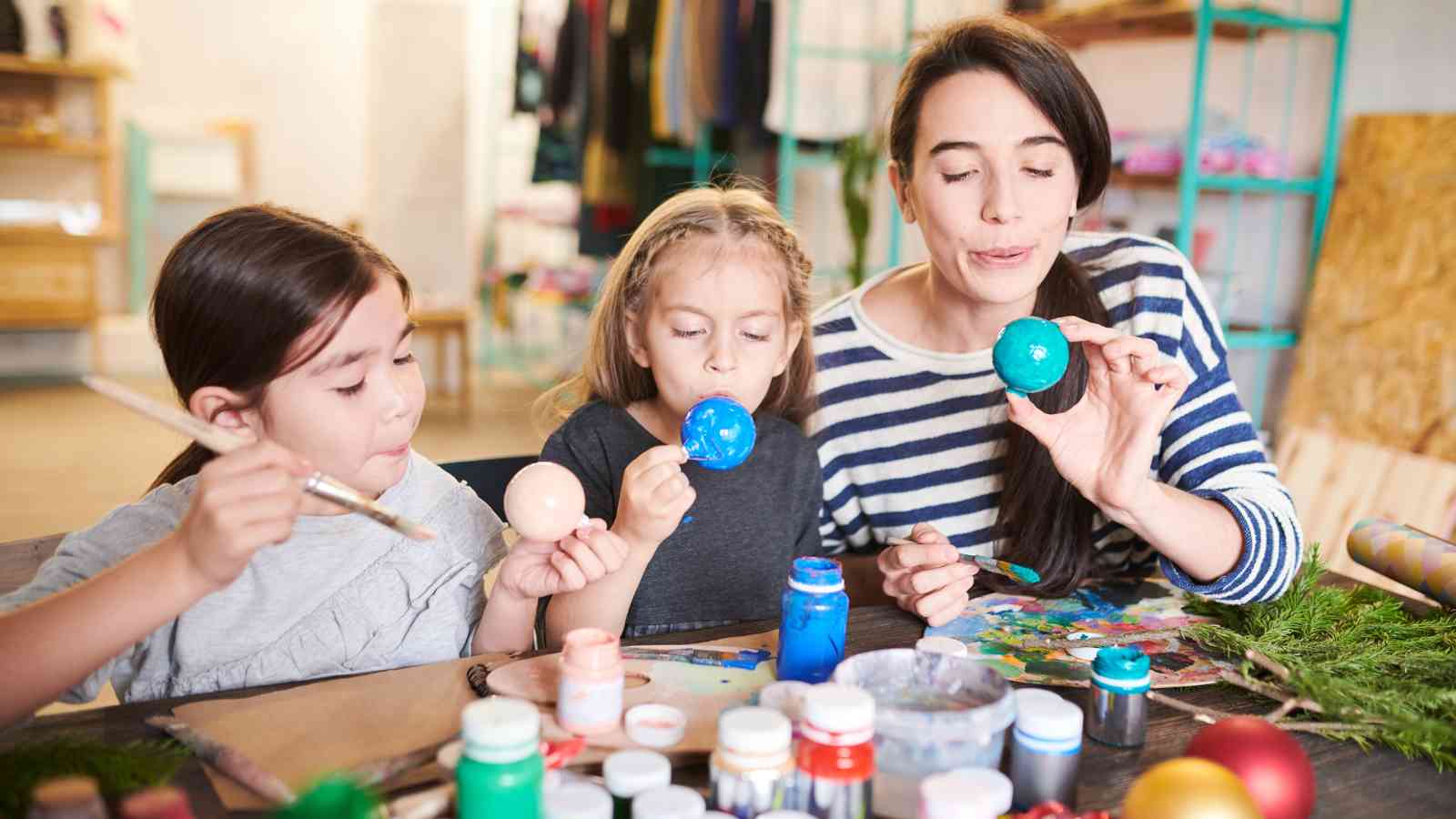 An adult and two children sit at a table painting eggs with brushes and paint, surrounded by art supplies.
